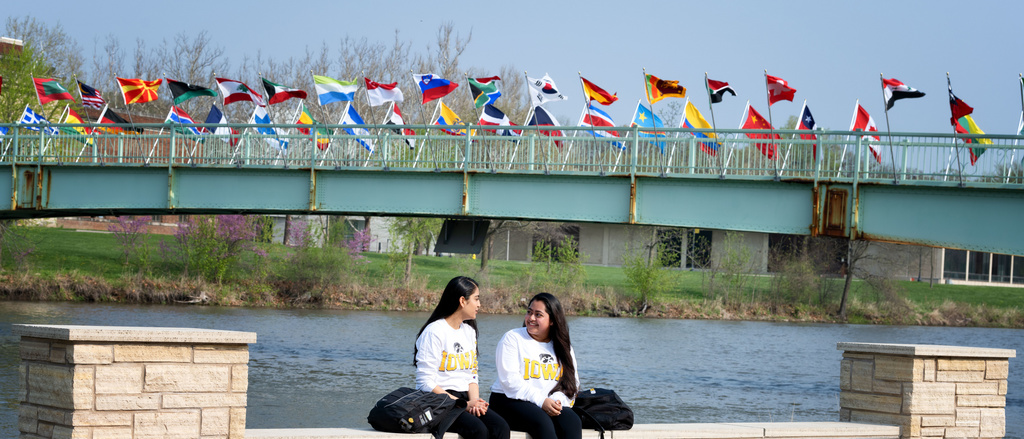 Two students sitting on a wall with the IMU bridge flags in the background.